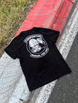A black ZeroPointOne t-shirt lies on a racetrack kerb with red, white, and black paint. The t-shirt features a bold white graphic of a cartoon character with a gear shift pattern on its forehead, holding a gear stick. Above the character, the text reads “ZEROPOINTONE” and below, “MANUAL FOR LIFE.” Grass, concrete, and asphalt make up the textured background.