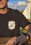 A black ZeroPointOne t-shirt on a man sitting in a beer garden. The t-shirt features a bold white graphic of a cartoon character with a gear shift pattern on its forehead, holding a gear stick. Above the character, the text reads “ZEROPOINTONE” and below, “MANUAL FOR LIFE.” Grass, concrete, and asphalt make up the textured background.