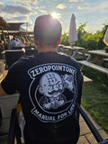 A black ZeroPointOne t-shirt on a man sitting in a beer garden. The t-shirt features a bold white graphic of a cartoon character with a gear shift pattern on its forehead, holding a gear stick. Above the character, the text reads “ZEROPOINTONE” and below, “MANUAL FOR LIFE.” Grass, concrete, and asphalt make up the textured background.